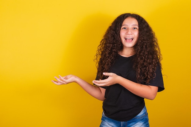 Brazilian latin american girl for afro hair yellow background smiling suggesting suggestion indication pointing with hands to the side negative space for advertising and publicity text recommending