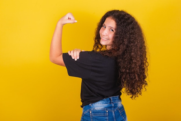 Brazilian latin american girl for afro hair yellow background\
smiling with hand on biceps happy indicating strength empowerment\
feliminism joyful confident optimistic