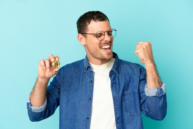 Foto uomo brasiliano che tiene un bitcoin su sfondo blu isolato che celebra una vittoria