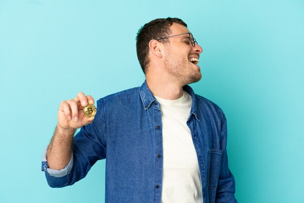 Brazilian man holding a Bitcoin over isolated blue background laughing in lateral position