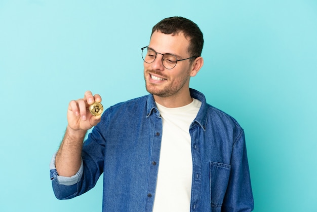 Brazilian man holding a Bitcoin over isolated blue background looking to the side and smiling