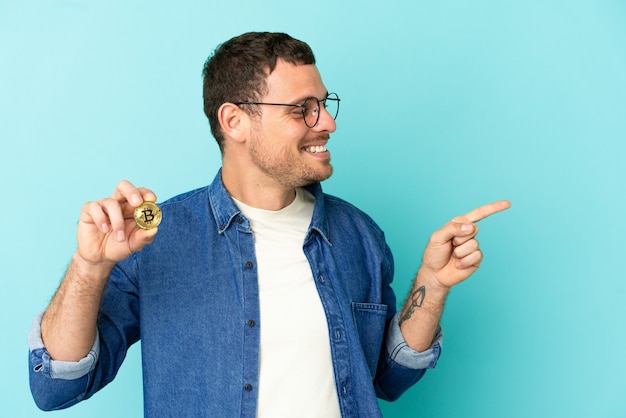 Brazilian man holding a Bitcoin over isolated blue background pointing finger to the side and presenting a product