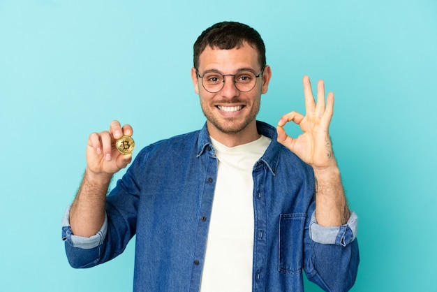 Brazilian man holding a Bitcoin over isolated blue background showing ok sign with fingers