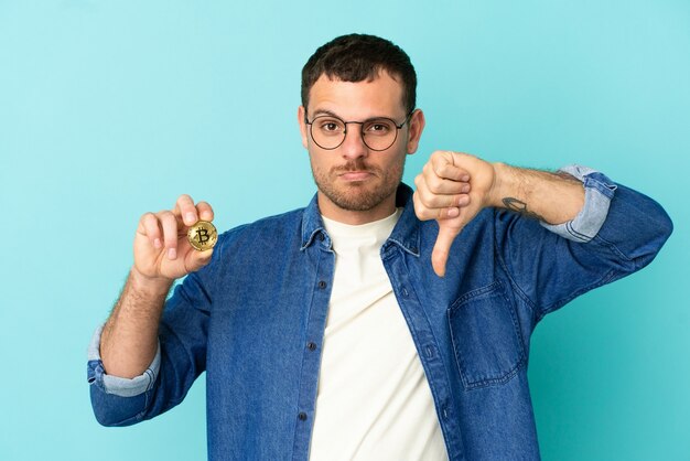 Brazilian man holding a Bitcoin over isolated blue background showing thumb down with negative expression