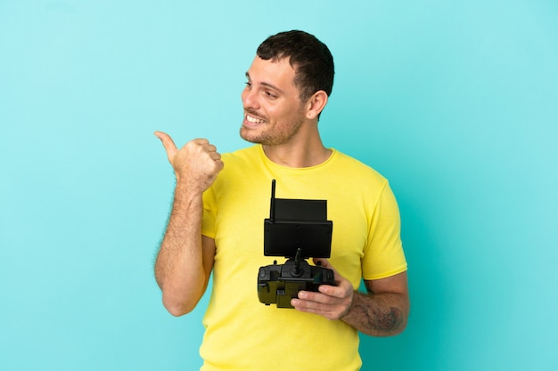 Brazilian man holding a drone remote control over isolated blue background pointing to the side to present a product