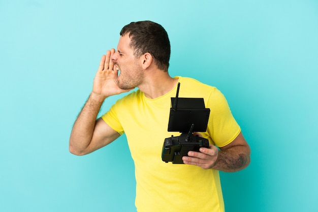 Brazilian man holding a drone remote control over isolated blue background shouting with mouth wide open to the side