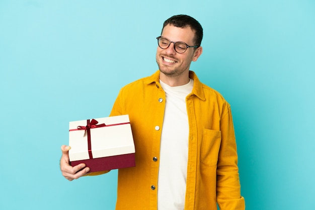 Uomo brasiliano che tiene un regalo su sfondo blu isolato guardando di lato e sorridente