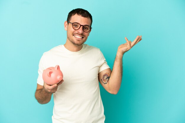 Brazilian man holding a piggybank over isolated blue background extending hands to the side for inviting to come