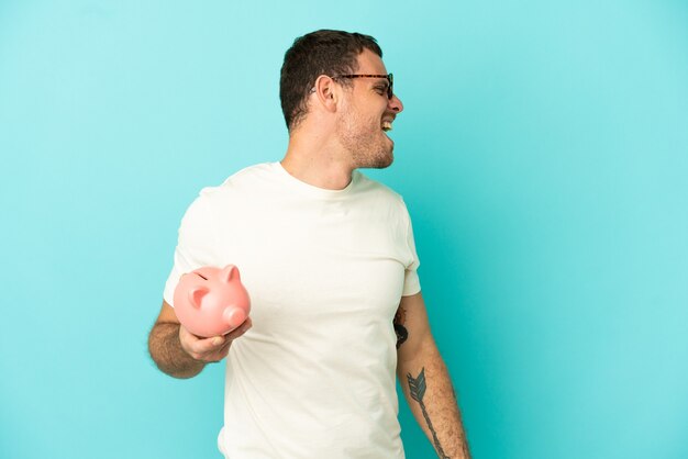 Brazilian man holding a piggybank over isolated blue background laughing in lateral position