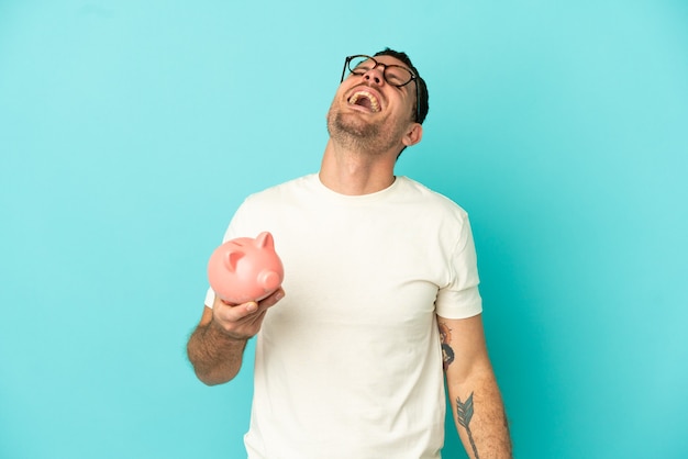 Brazilian man holding a piggybank over isolated blue background laughing