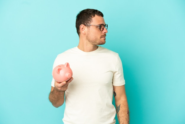 Brazilian man holding a piggybank over isolated blue background looking to the side