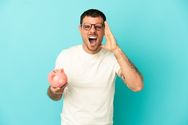 Brazilian man holding a piggybank over isolated blue background shouting with mouth wide open