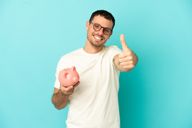 Brazilian man holding a piggybank over isolated blue background with thumbs up because something good has happened