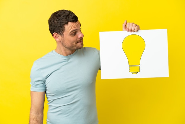 Brazilian man over isolated purple background holding a placard with bulb icon