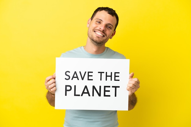 Бразильский мужчина на изолированном фиолетовом фоне держит плакат с текстом «Спасите планету» со счастливым выражением лица