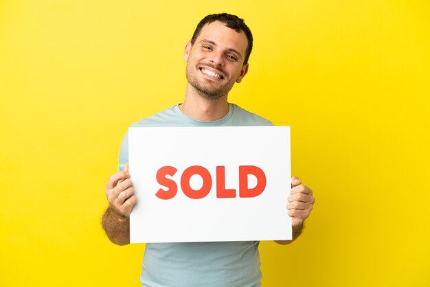 Brazilian man over isolated purple background holding a placard with text SOLD with happy expression