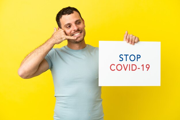 고립된 보라색 배경 위에 있는 브라질 남성은 Stop Covid 19라는 문구가 적힌 플래카드를 들고 전화 제스처를 하고 있습니다.