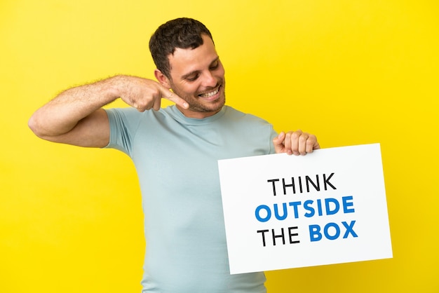 Бразильский мужчина на изолированном фиолетовом фоне держит плакат с текстом Think Outside The Box и указывает на него