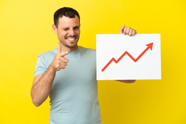 Brazilian man over isolated purple background holding a sign with a growing statistics arrow symbol and pointing to the front