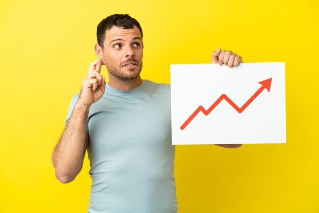 Brazilian man over isolated purple background holding a sign with a growing statistics arrow symbol with fingers crossing