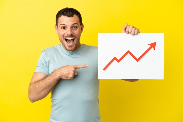 Brazilian man over isolated purple background holding a sign with a growing statistics arrow symbol with surprised expression