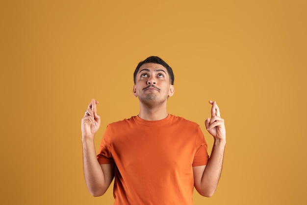 Brazilian man in orange tshirt making cross fingers gesture and looking up yellow background studio