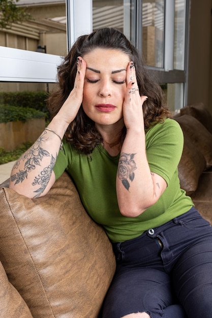 Brazilian tattooed and worried with both hands on her head temple sitting on the sofavertical