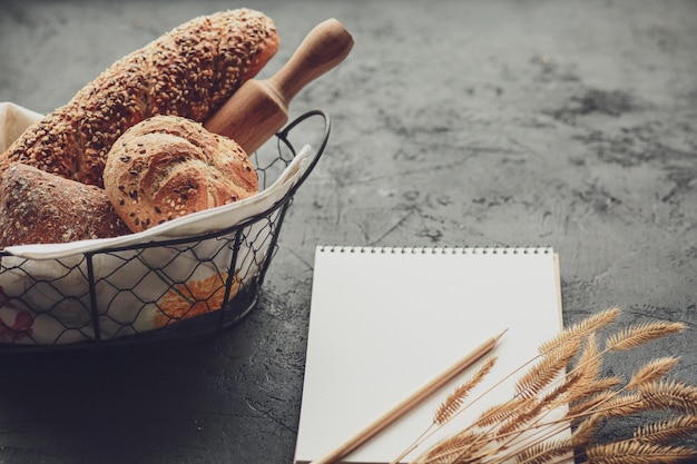 Bread in a basket on a black surface. Assorted bread in a metal basket. Place for recipe and text. Bake bread with a rolling pin and flour. Rye bread, buckwheat rolls and baguette with seeds.