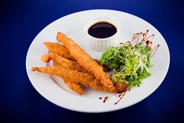 Photo breaded chicken strips with sauce and lettuce leaves. flat lay top view on a white plate on a blue background.