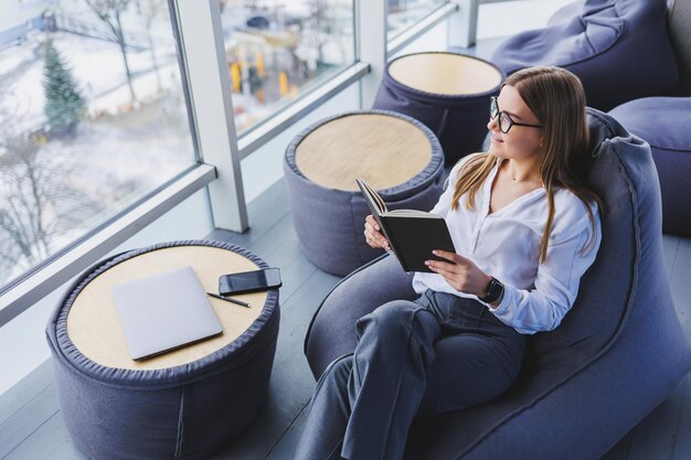 Break Smiling young woman in glasses relaxing on bean bag chair sitting at table and relaxing using notepad happy woman leaning back enjoying work feeling satisfied