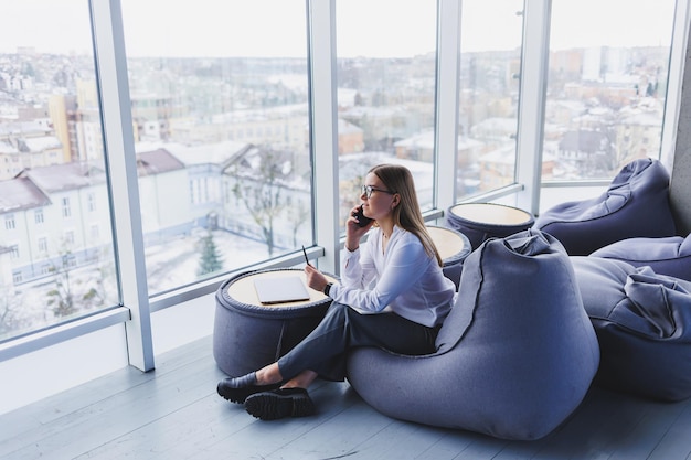 Break Smiling young woman in glasses relaxing on bean bag chair sitting at table and relaxing using notepad happy woman leaning back enjoying work feeling satisfied