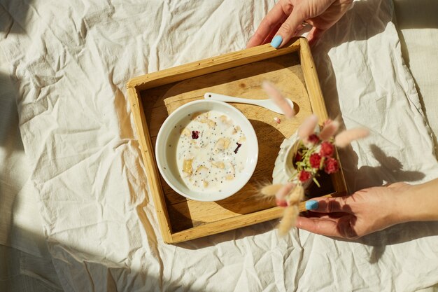 Breakfast in bed, female hands put try with bowl muesli, granola and flower in sunlight at home, chambermaid bringing tray with breakfast in hotel room, good service