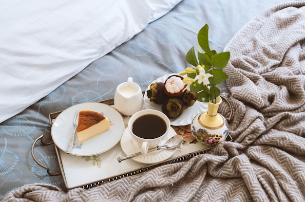 Breakfast in bed slice of cheesescake and fruit with cup of\
coffee and flower in a tray.