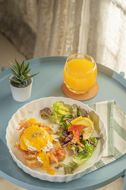 A colazione o brunch, le uova alla benedict servono con pancetta fritta e pane tostato e insalata di verdure per contorno in un piatto bianco o piatto su un panno bianco con striscia verde e vengono con succo d'arancia