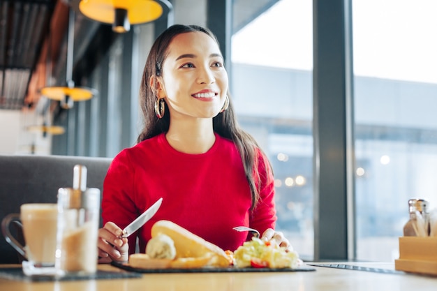 Breakfast in cafe. Close up of beaming appealing woman wearing red sweater eating breakfast in cafe