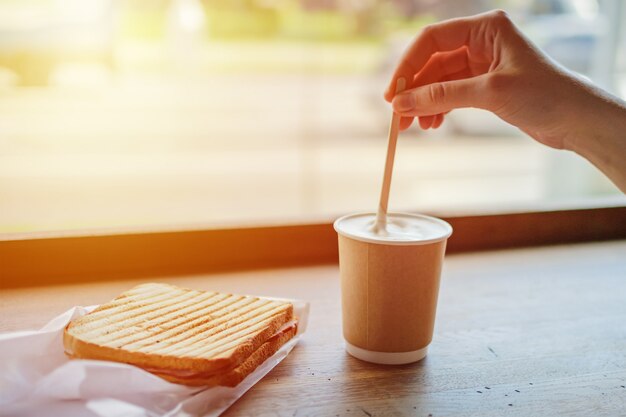 Breakfast in cafe with coffee and toast. Woman's hand stirs coffee in paper cup