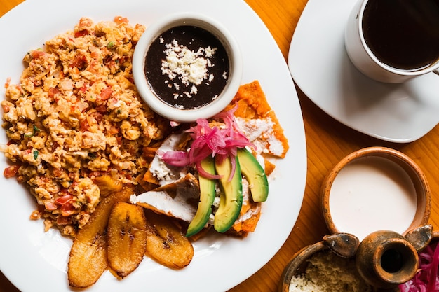 Photo breakfast of chilaquiles with beans, avocado, onion, egg, fried banana, cream and coffee. food.