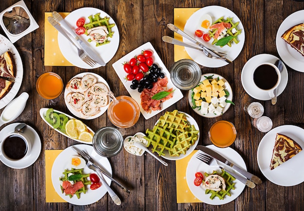 Breakfast food table. Festive brunch set, meal variety with spinach waffles, salmon, cheese, olives, chicken rolls and cheesecake. Top view. Flat lay