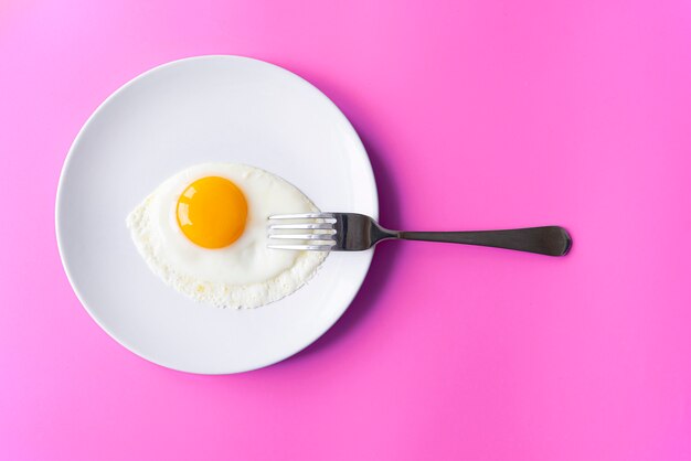 Breakfast, fried egg, yolk, omelet and fork on white plate with color table, creative picture