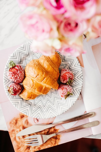 Breakfast from the croissant on the table with a fork and knife and a colorful bouquet