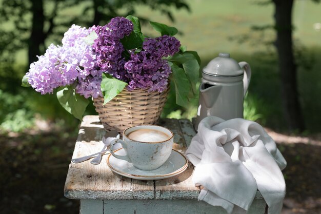 Breakfast in the garden: eclairs, cup of coffee, coffee pot, lilac flowers in a basket.