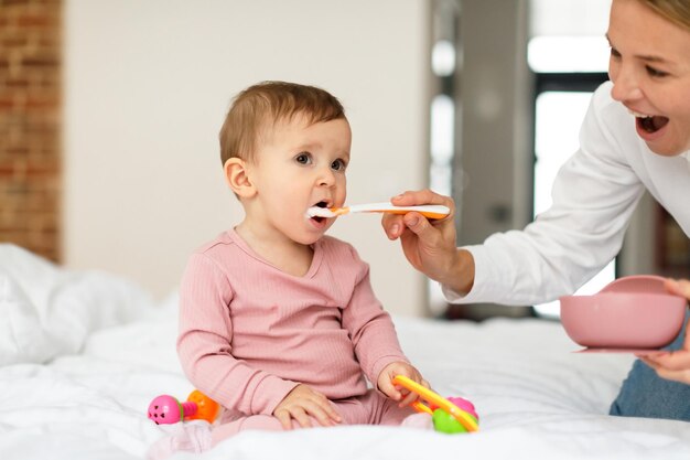 Breakfast for kid Mother feeding her adorable baby daughter with porridge sitting on bed in bedroom interior free space