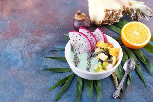 Breakfast kiwi smoothie bowl topped with pitaya, pineapple, chia seeds and berries with palm leaf on stone background, top view