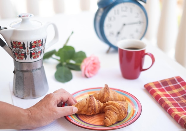 テラスの外の朝の朝食 人間の手はクロワッサンを持ってコーヒーとバラを手に取り、一日を明るくします ポジティブな瞬間 大きな古い目覚まし時計