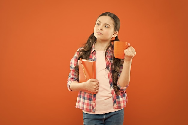 Breakfast the most important meal of the day Little child having breakfast on orange background Cute pupil holding book and drink for breakfast Small girl thinking during her school breakfast