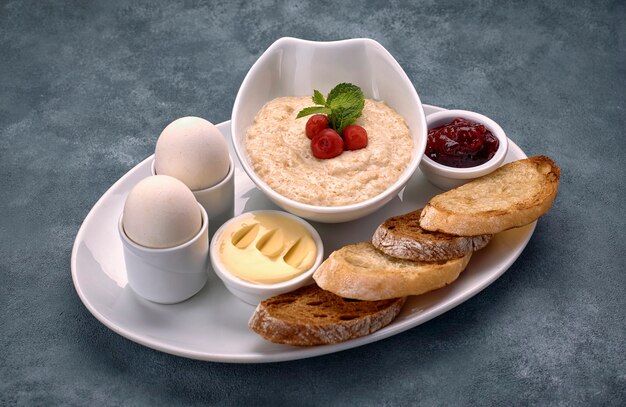 Colazione a base di porridge, burro, uova sode, marmellata e pane tostato, crostini di pane, su una superficie grigio-blu