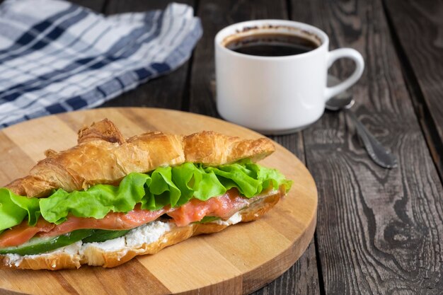 Breakfast sandwich croissant with red fish salmon cottage cheese cucumbers and salad on a dark wooden background with coffee and napkin