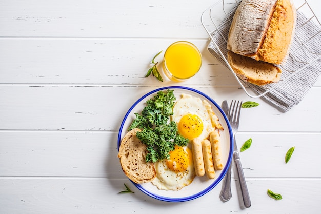 La colazione viene servita con uova fritte, insalata, muffin e succo d'arancia sul tavolo di legno bianco.