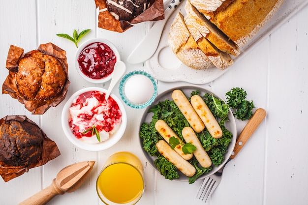 Breakfast served with muffins, grilled sausages, juice, fresh bread and parfait on white wooden table.