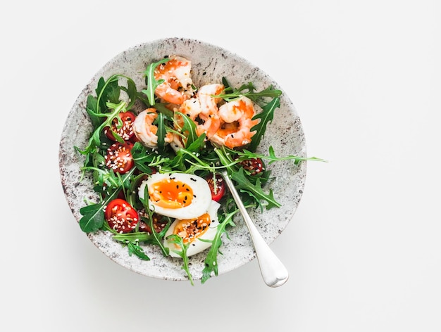 Breakfast snack bowl arugula cherry tomatoes salad with boiled egg and fried shrimp on a light background top view